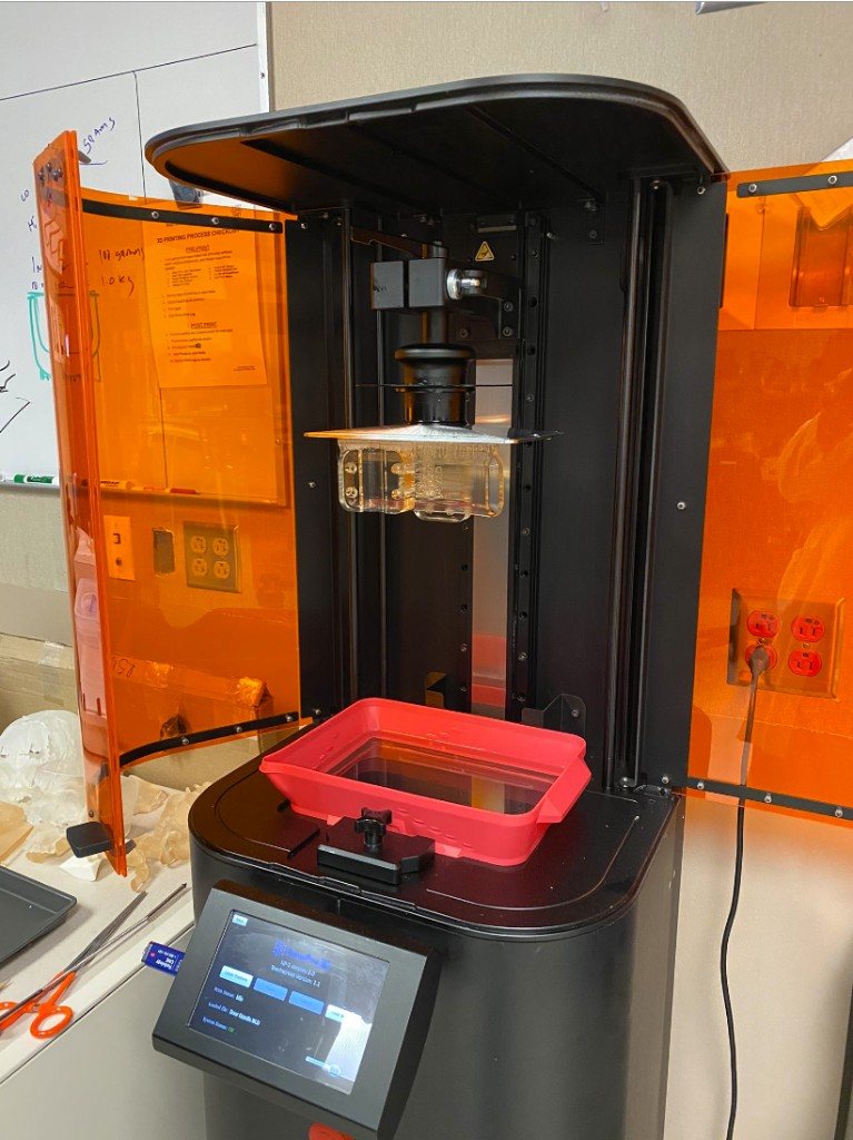 SLA 3D printer in a lab with orange UV doors open, a clear printed part on the build plate, and anatomical models on the bench.